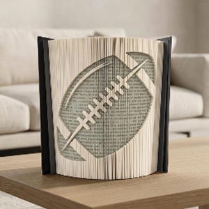 Op de afbeelding: Decoratieve boeksculptuur met een football-ontwerp uitgesneden in de pagina's. Het open boek onthult de vorm van de football, met zwarte kaften. Een uniek woondecoratie-item. Ongeveer 20 cm hoog.