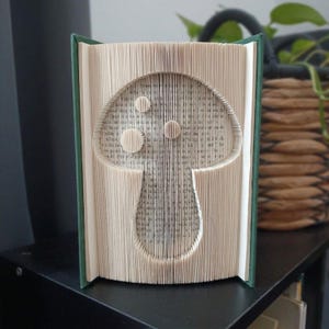 May include: A book sculpture featuring folded pages that form the shape of a mushroom with a rounded cap and a stem. The book has a dark green cover and the interior pages are cream-coloured with visible black printed text throughout the folds.