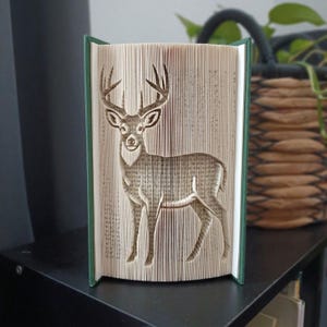 May include: A book sculpture featuring a folded page design that creates the silhouette of a standing deer with antlers. The book has a dark green cover and the pages are vertically aligned to form the detailed shape of the animal's body.