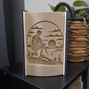 Könnte beinhalten: Eine Buchfaltkunst-Skulptur, die die Silhouette eines Soldaten auf felsigem Gelände zeigt. Das Design entsteht durch präzises Falten der Buchseiten, wodurch eine Szene mit Sonne, Wolken und Landschaftselementen aus beigefarbenem Papier sichtbar wird.