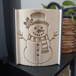 Può includere: Una scultura realizzata con un libro piegato che raffigura un pupazzo di neve, creata piegando con precisione le pagine. Il pupazzo di neve indossa un cilindro con un rametto di agrifoglio, una sciarpa e ha braccia a forma di ramoscelli. Pagine color crema.