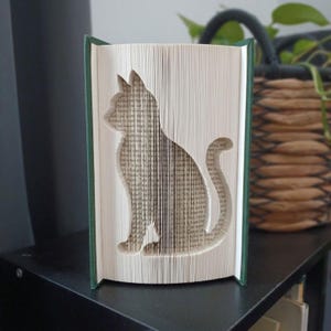 May include: A folded book art sculpture featuring a cat silhouette created by precisely folding the individual pages of a hardcover book. The book has a dark green spine and the pages are arranged to form the distinct shape of a seated cat.