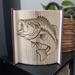 Peut inclure: Une sculpture en livre avec un design de pages pliées formant la silhouette d'un poisson bar. Les pages sont précisément marquées et pliées pour créer le contour détaillé du poisson, incluant ses nageoires, ses écailles et sa bouche ouverte.