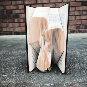 May include: A book with pages folded into the shape of a mushroom. The book is open and the pages are fanned out to create the mushroom shape. The book is sitting on a brick surface.