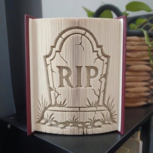 Peut inclure: Une sculpture en livre plié représentant une pierre tombale avec les lettres RIP clairement visibles au centre. Les pages sont pliées avec précision pour former une stèle fissurée avec de l'herbe à la base et une couverture de livre rouge foncé.