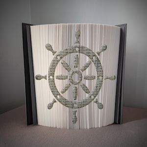 May include: A decorative book with pages folded to create a ship's wheel design. The wheel is gray with a textured appearance, set against the white pages of the book. The book has a dark gray spine and cover.
