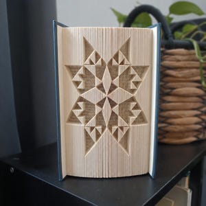 May include: A book sculpture featuring a star pattern created by precisely folding the pages. The book has a dark blue cover and the interior pages are cream-coloured, showing text and geometric shapes formed by the folded paper edges.