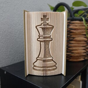 May include: A book sculpture featuring a folded page design that forms the shape of a chess king piece. The pages are cream-coloured with visible printed text, and the sculpture is standing upright with the spine facing away from the viewer.