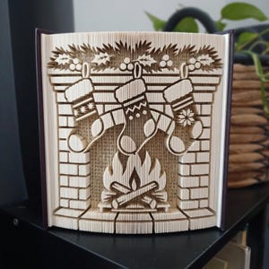 May include: A folded book art sculpture featuring a fireplace scene with three hanging stockings, a brick mantel, and a burning fire with logs. The pages are precisely folded to create a three-dimensional relief of a holiday hearth.
