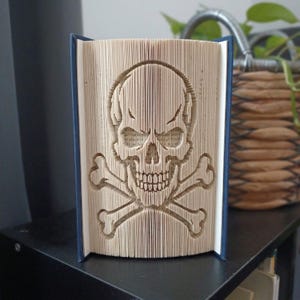 Op de afbeelding: Een gevouwen boekkunstsculptuur met een schedel- en gekruiste beenderenontwerp, gemaakt door de individuele pagina's van een hardcover boek nauwkeurig te vouwen. De pagina's zijn crèmekleurig en de donkerblauwe kaft is aan de zijkanten zichtbaar.