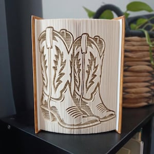 May include: A folded book art sculpture featuring a pair of cowboy boots created by precisely folding the pages of a hardback book. The cream-coloured paper pages are manipulated to form the distinct silhouette and decorative stitching of boots.
