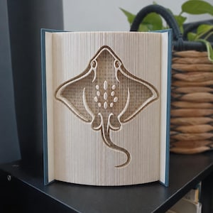 Peut inclure: Une sculpture de livre avec un design de pages pliées formant la silhouette d'une raie. Les pages sont précisément pliées pour dessiner les contours de la créature, incluant ses larges nageoires, sa longue queue et des motifs ovales.