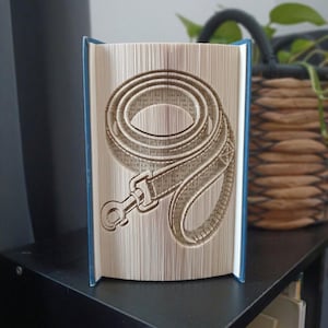 Puede incluir: Una escultura de libro plegado que presenta un diseño detallado de una correa para perro enrollada con un clip de metal. Las páginas están dobladas y marcadas con precisión para crear la forma de la correa, con las cubiertas azules visibles.