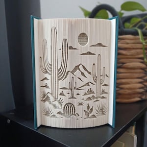 May include: A folded book art sculpture featuring a desert landscape with saguaro cacti, a barrel cactus, and prickly pear cacti. The pages are intricately folded to create a three-dimensional scene of mountains, clouds, and a sun in the background.