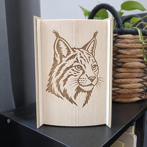 Könnte beinhalten: Eine Buchfaltkunst-Skulptur mit einem detaillierten Luchs-Kopf-Design, das durch präzises Falten der Buchseiten entsteht. Die hellen Papierkanten bilden die Silhouette und die Gesichtszüge des Wildkatzen-Motivs mit feinen Texturen.