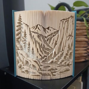 May include: A folded book art sculpture featuring a mountain landscape with pine trees and rocky cliffs. The pages are precisely folded to create a three-dimensional relief of a valley scene, with the blue book cover visible on the sides.