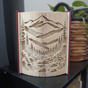 Op de afbeelding: Een boeksculptuur met ingewikkeld gevouwen pagina's die een landschap vormen. Het ontwerp toont een bergketen aan de bovenkant, met lagen dennenbomen en rotsachtig terrein eronder. Het boek heeft een donkerrode kaft die aan de zijkanten zichtbaar is.