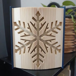 May include: A folded book art sculpture featuring a snowflake design created by precisely folding the individual pages of a book. The cream-colored paper pages are arranged to form a symmetrical, six-pointed snowflake pattern with dark blue covers.