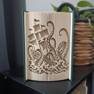 Op de afbeelding: Een boeksculptuur met gevouwen pagina's die een driedimensionaal beeld vormen van een zeilschip dat wordt aangevallen door een grote octopus met lange tentakels die uit gestileerde golven oprijzen. Het boek heeft een donkergroene kaft.
