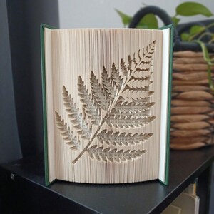 May include: A book sculpture featuring pages folded to create a detailed fern leaf pattern. The book has a dark green cover and stands upright, displaying the intricate paper art formed by the precisely folded edges of the printed text pages.