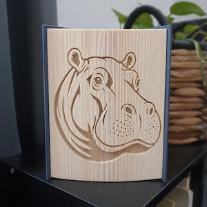 Może przedstawiać: Rzeźba wykonana z zagiętych stron książki, przedstawiająca szczegółowy, trójwymiarowy relief głowy hipopotama. Strony są precyzyjnie pozaginane, aby stworzyć wizerunek zwierzęcia, z widoczną fakturą krawędzi papieru na całej powierzchni.