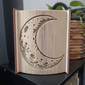 May include: A folded book sculpture featuring a crescent moon design created by precisely folding the individual pages of a hardcover book. The cream-colored paper edges form the detailed shape of the moon with visible craters and textures.