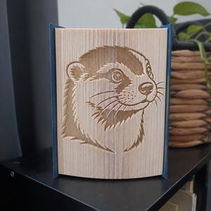 May include: A folded book art sculpture featuring a detailed profile of an otter face created by precisely folding the pages of a book. The beige paper edges form the image of the animal, while the dark blue book covers are visible on the sides.