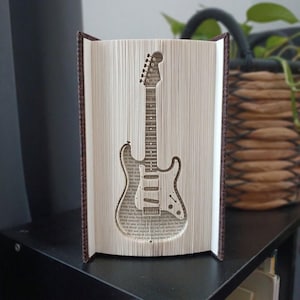 Pode incluir: Uma escultura de livro com um design de páginas dobradas que cria a silhueta de uma guitarra elétrica. As páginas são de cor creme com texto preto visível e o livro tem capas castanho-escuras posicionadas verticalmente.