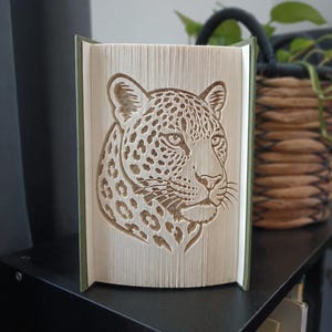 May include: A folded book art sculpture featuring a detailed leopard head design created by precisely folding the pages of a book. The sculpture has a green spine and shows the leopard's face, ears, and whiskers in the white paper edges.
