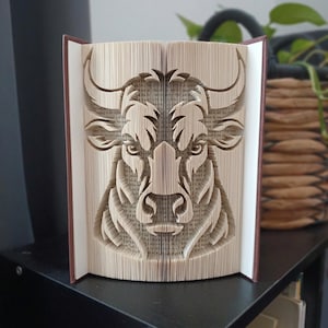 Peut inclure: Une sculpture de livre représentant une tête de taureau créée en pliant les pages d'un livre à couverture rigide. Les pages en papier couleur crème sont précisément manipulées pour former les traits du visage, les cornes et les oreilles.
