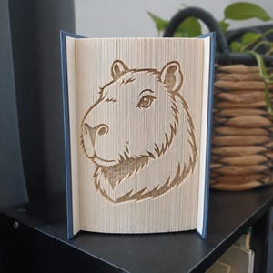 Peut inclure: Une sculpture de livre avec un design de pages pliées formant le profil détaillé d'une tête de capybara. Le livre possède une couverture bleue et les pages sont disposées pour créer la forme du visage, des oreilles et la texture du pelage.