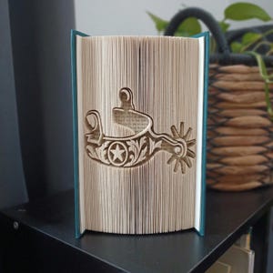 Peut inclure: Une sculpture de livre avec un design de pages pliées formant la silhouette d'une éperon western. Les pages sont de couleur crème et le livre possède une couverture bleu sarcelle. Le motif de l'éperon inclut une étoile et des volutes.