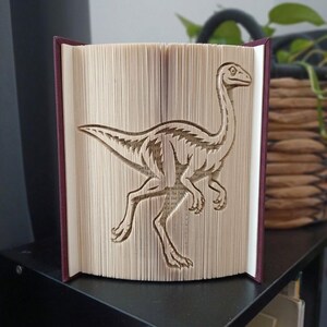 Op de afbeelding: Een gevouwen boekkunstsculptuur met een gedetailleerd silhouet van een dinosaurus, gemaakt door de individuele pagina's van een hardcover boek nauwkeurig te vouwen. Het boek heeft donkerrode kaften en crèmekleurige pagina's met tekst.