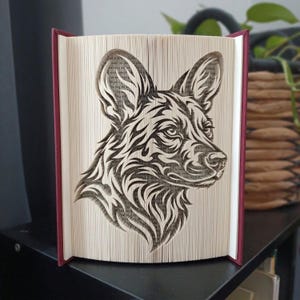 Op de afbeelding: Een gevouwen boekkunstsculptuur met het gedetailleerde profiel van een wilde hond. De pagina's zijn nauwkeurig gevouwen om de kop en oren van het dier te vormen, waarbij de donkere tekst op het papier contrasteert met de witte randen.
