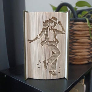 May include: A book sculpture featuring folded pages that create a silhouette of a person in a suit and hat performing a dance move. The pages are cream-coloured with visible black printed text, forming the shape of a figure in a dynamic pose.
