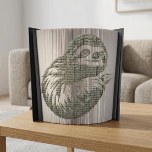 May include: A book sculpture featuring a sloth design created by folding the pages. The sloth image is in green and grey tones. The book has a black spine and is displayed on a wooden table. The book is approximately 20 cm tall.