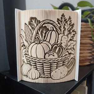 Könnte beinhalten: Eine Buchfaltkunst-Skulptur mit einem detaillierten Relief eines geflochtenen Korbs, gefüllt mit verschiedenen Gemüsesorten wie Kürbissen, Mais, Karotten und Blattgemüse. Die Seiten sind präzise gefaltet, um eine dreidimensionale Ernteszene zu erzeugen.