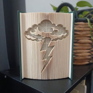 Peut inclure: Une sculpture de livre avec des pages pliées formant la silhouette d'un nuage avec un éclair descendant. Le livre possède une couverture vert foncé et les pages sont de couleur crème avec du texte imprimé noir visible sur l'ensemble.