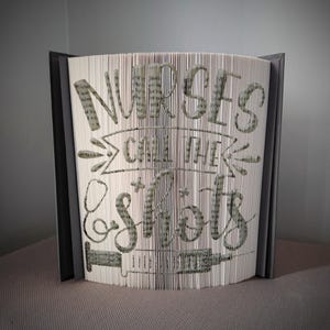 May include: A handmade book sculpture with the phrase "Nurses Call the Shots" cut into the pages. The text is in a handwritten style, with a syringe and stethoscope design. The book has a grey cover.