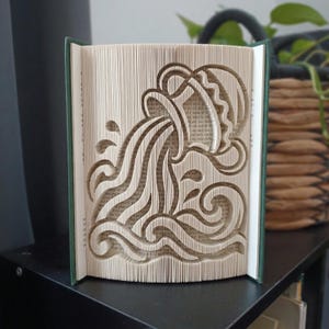 Op de afbeelding: Een gevouwen boekkunstsculptuur met het sterrenbeeld Waterman. De pagina's zijn nauwkeurig gevouwen om de vorm van een waterkruik te creëren die vloeistof in golven giet. Het boek heeft een donkergroene kaft en crèmekleurige pagina's.