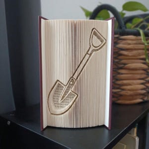 Könnte beinhalten: Eine Buchskulptur mit gefalteten Seiten, die eine klare, dreidimensionale Silhouette einer Schaufel bilden. Das Buch hat dunkelweinrote Einbände und die Innenseiten sind präzise geknickt, um den Stiel und das Blatt des Werkzeugs zu formen.