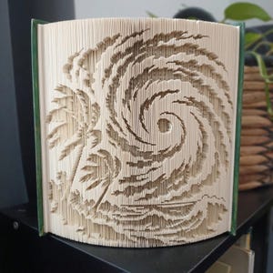 Op de afbeelding: Een gevouwen boekkunstsculptuur met een gedetailleerd, driedimensionaal orkaan- of cycloonpatroon, gemaakt door de individuele pagina's van een hardcover boek nauwkeurig te vouwen. De crèmekleurige paginaranden vormen een wervelend ontwerp.