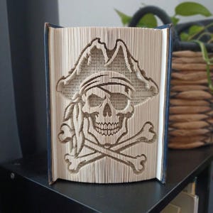 Peut inclure: Une sculpture en livre plié représentant un crâne de pirate détaillé avec des os croisés. Les pages du livre sont précisément pliées et marquées pour créer l'image en trois dimensions d'un crâne portant un bandana et un chapeau de pirate.