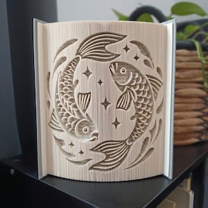 May include: A book sculpture featuring intricate folded pages that form a circular design of two fish swimming in a yin-yang pattern, accented with small four-pointed star shapes. The paper edges are precisely folded to create the fish imagery.