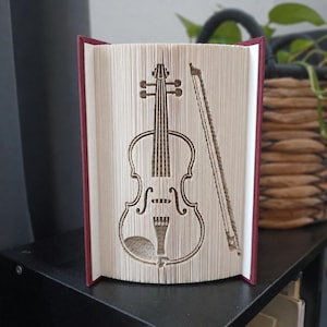 Op de afbeelding: Een gevouwen boekkunstsculptuur met een gedetailleerd ontwerp van een viool en strijkstok, gemaakt door de pagina's van een boek met een donkerrode kaft nauwkeurig te vouwen. De crèmekleurige paginaranden vormen het silhouet.