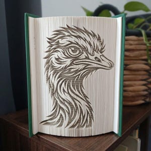 May include: A book sculpture featuring a folded page art design of an emu head in profile. The pages are precisely creased and folded to create the detailed silhouette of the bird, with the dark text on the paper forming the emu's features.