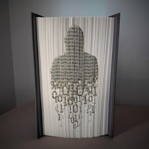 Puede incluir: Un libro abierto con la silueta de una persona recortada en las páginas. La figura está llena de texto y código binario. El libro tiene una cubierta gris oscuro. Las páginas están cortadas para crear la forma de una persona.