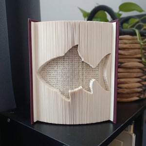 Puede incluir: Una escultura de libro con páginas dobladas que crean la silueta de un tiburón. El libro tiene una cubierta de color púrpura oscuro y páginas de papel blanco. Las páginas están dobladas con precisión para formar la silueta del pez.