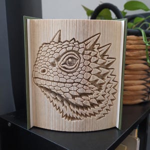 May include: A book sculpture featuring a folded page art design of a bearded dragon head in profile. The pages are precisely creased to create the textured, scaled appearance of the reptile, including its eye, spikes, and distinct facial features.