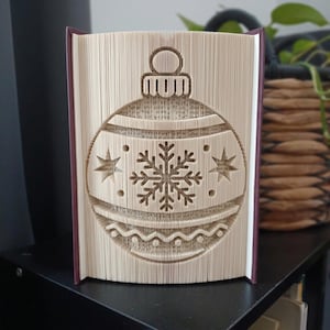 Könnte beinhalten: Eine gefaltete Buchkunst-Skulptur mit einem Weihnachtskugel-Motiv. Die Seiten sind präzise geknickt, um eine zentrale Schneeflocke, zwei seitliche Sterne und eine dekorative Wellenlinie am unteren Rand in einer runden Form zu bilden.